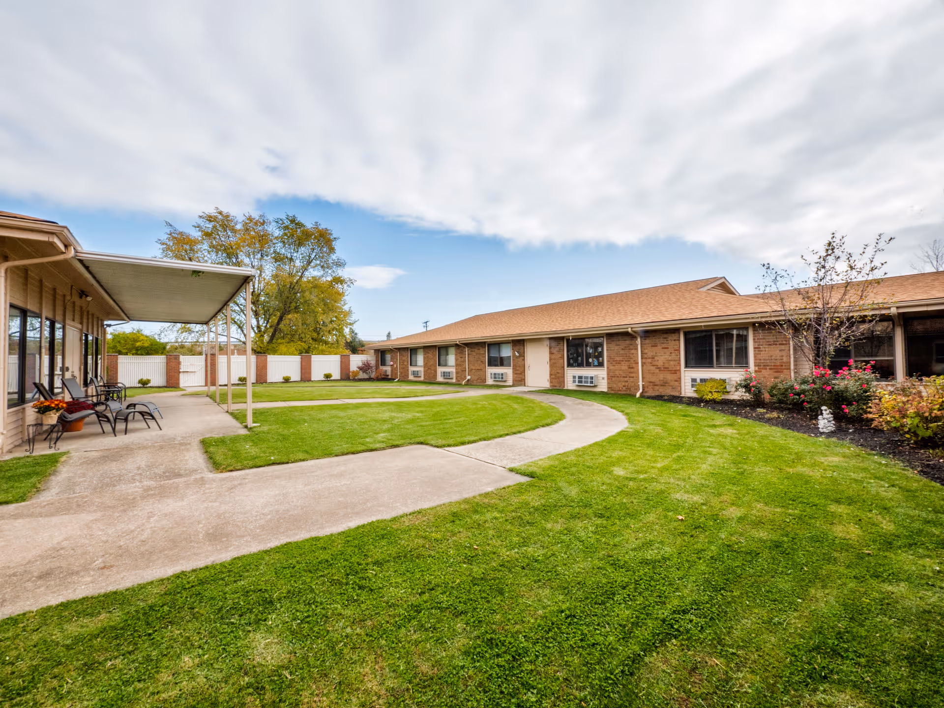 Outdoor courtyard area of a senior living facility with a curved concrete walkway, green grass, and a covered patio with chairs. The building is single-story with brick walls and multiple windows. There are some bushes and small trees along the building.
