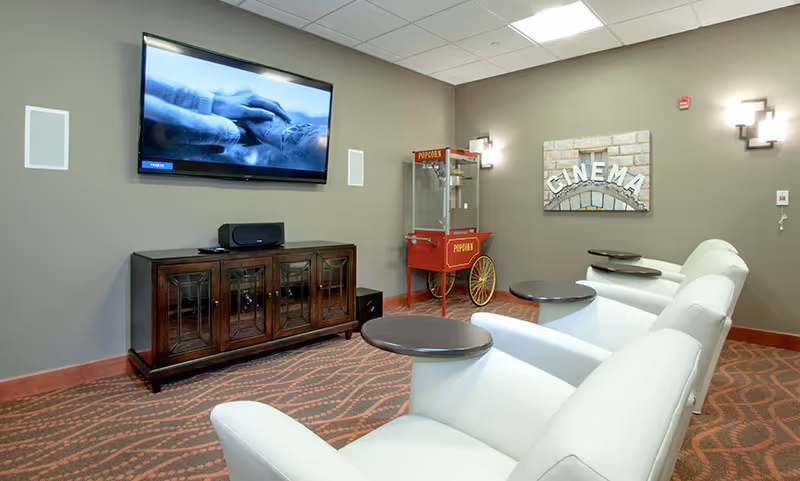 A cozy cinema room with three white recliner chairs each equipped with a small round table. A large flat-screen TV is mounted on the wall above a wooden cabinet. To the right of the TV, there is a vintage-style red popcorn machine and a wall decoration with the word 'CINEMA'. The room has patterned carpet flooring and soft wall lighting.