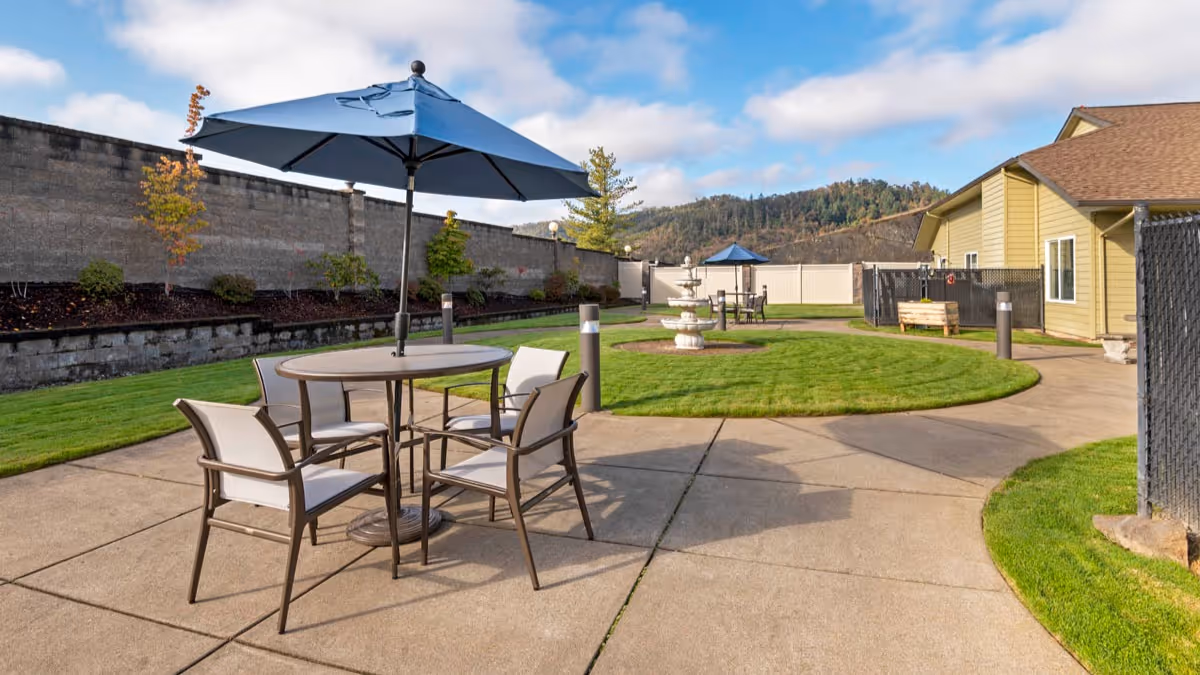 Outdoor patio area with a round table and four chairs under a blue umbrella, surrounded by a concrete walkway and green grass. There is a multi-tiered white fountain in the background, a yellow building to the right, and a tall stone wall with some small trees and shrubs along it. Hills and a partly cloudy sky are visible in the distance.