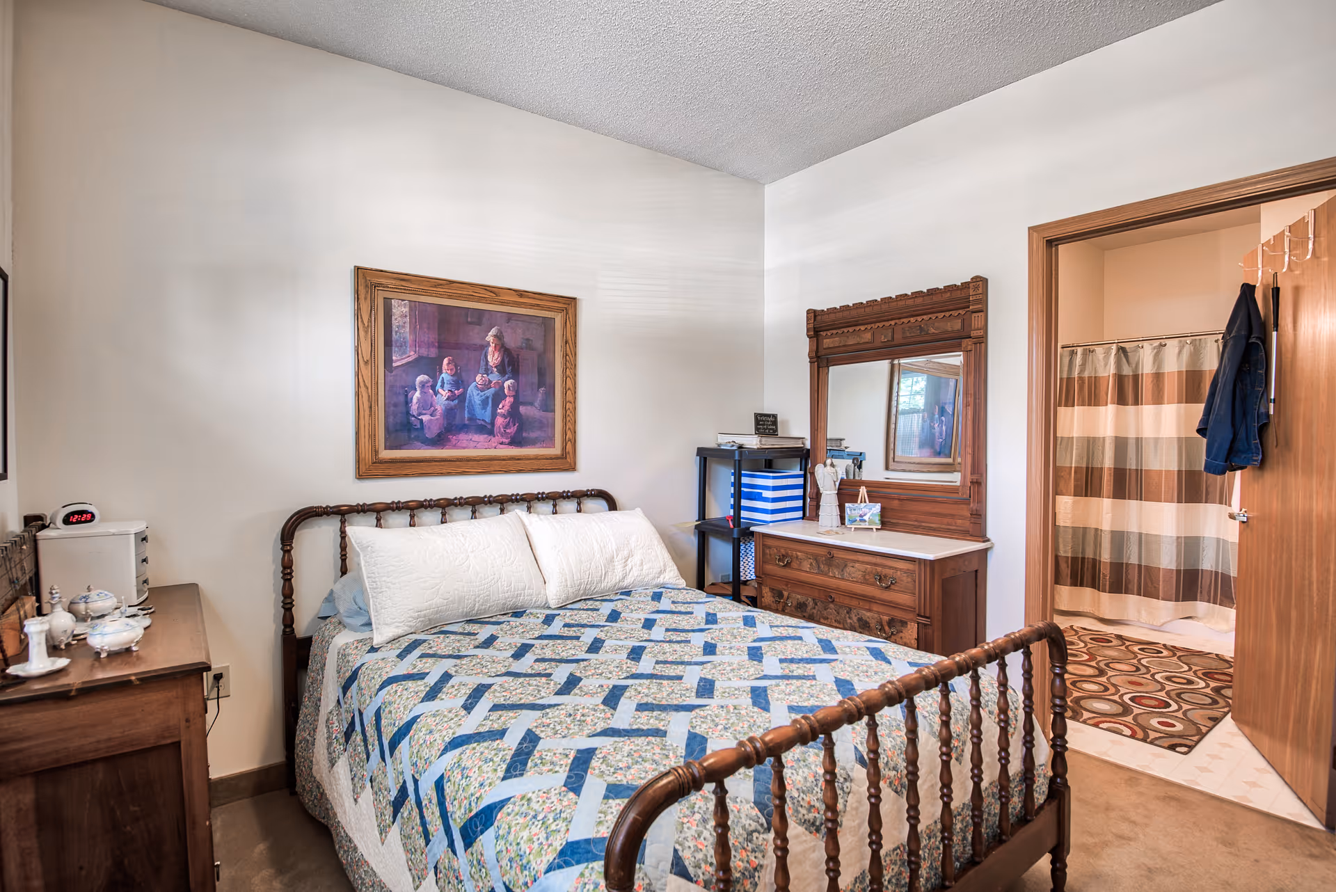 A cozy bedroom with a wooden bed frame and a quilted bedspread in blue and floral patterns. There is a wooden dresser with a large mirror and a small shelving unit with storage boxes. A framed painting of a woman and children hangs on the wall above the bed. An open door reveals a bathroom with a striped shower curtain and a circular patterned rug on the floor.