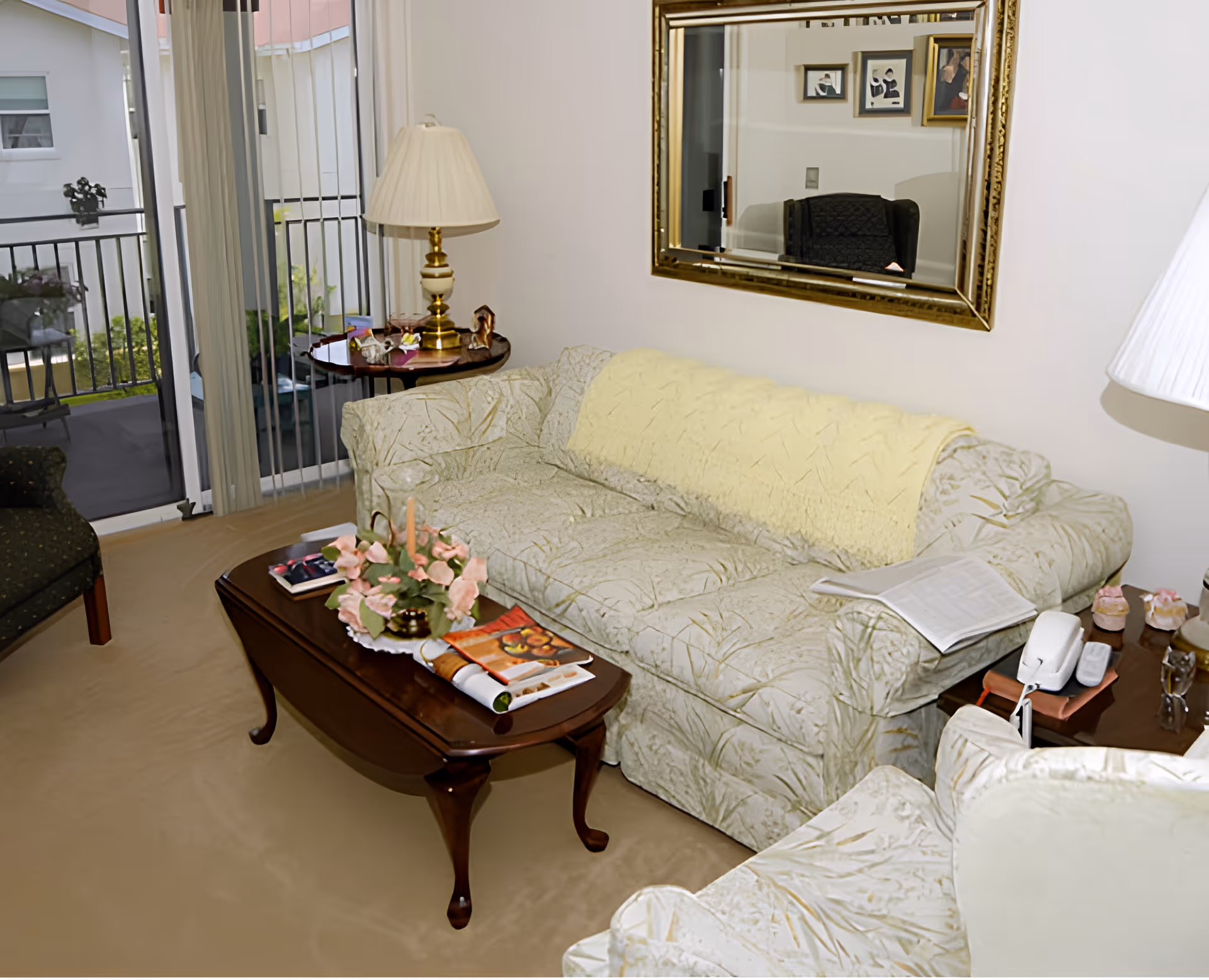 Cozy living room with a floral-patterned sofa, coffee table with flowers and magazines, side tables and lamps, and sliding glass doors to a balcony.