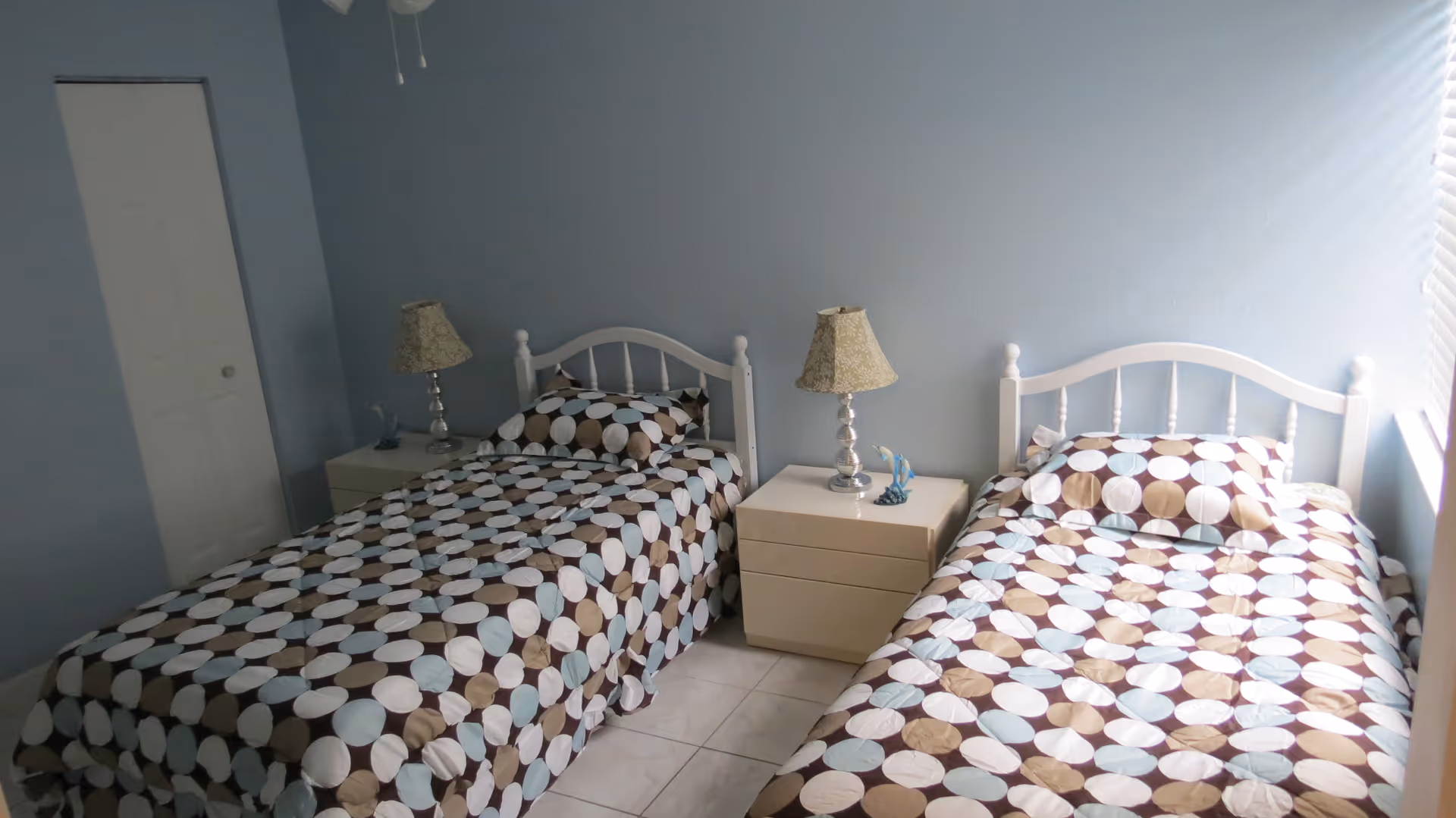 A bedroom with two single beds placed side by side, each with a white headboard and matching polka dot bedspreads in shades of brown, blue, and white. Between the beds is a small beige nightstand with two drawers, holding two decorative lamps and a small blue figurine. The walls are painted light blue, and there is a closed white door on the left side of the room. Natural light is coming in from a window on the right.