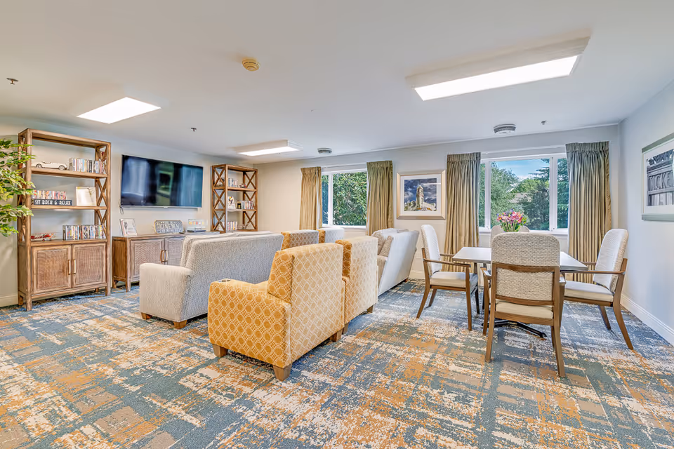 Bright senior living common room with sofas and armchairs facing a wall-mounted TV, bookshelves, and a dining table by windows.