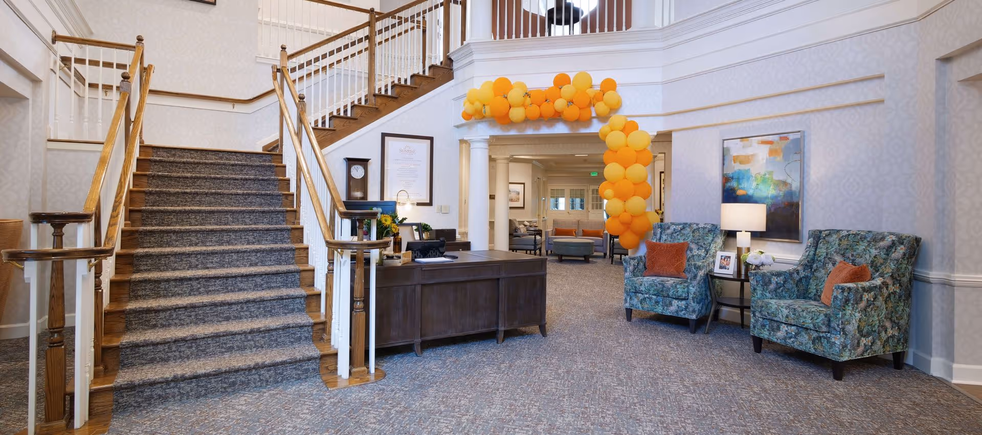 Interior view of a senior living facility lobby with a carpeted staircase with wooden handrails on the left, a dark wooden reception desk in the center, and two floral upholstered armchairs with orange pillows on the right. Above the entrance to another room is an arch decorated with yellow and orange balloons. The walls are light-colored with subtle patterns, and there is a colorful abstract painting and a table lamp near the armchairs.