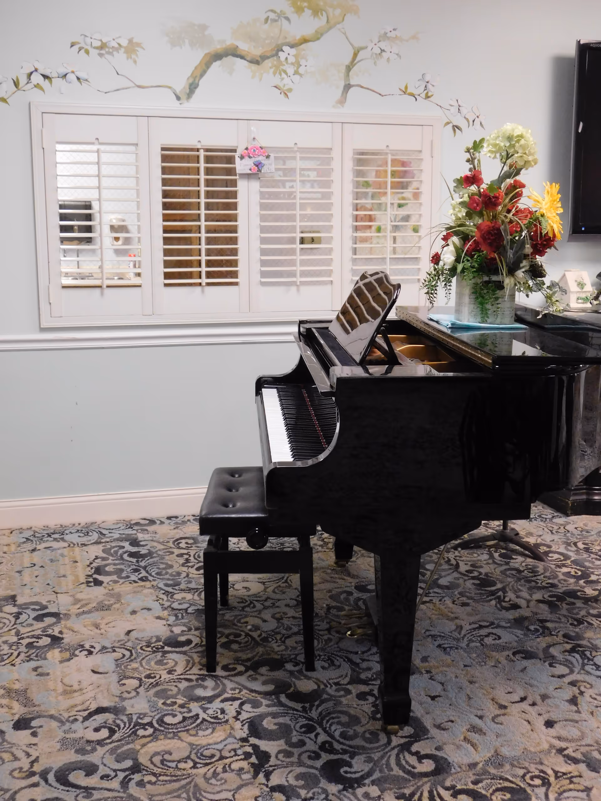 A black grand piano with a matching bench sits on a patterned carpet in a room. Behind the piano is a wall with white window shutters and a floral mural above. To the right of the piano is a vase with a colorful bouquet of flowers.