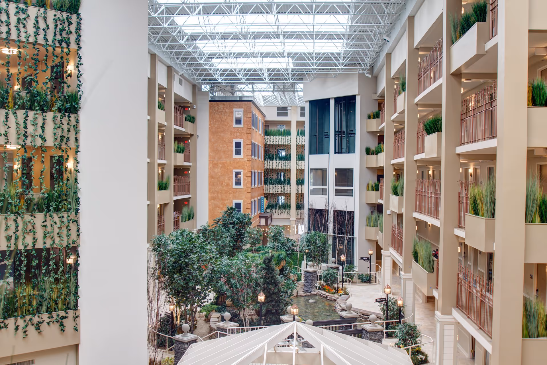 Indoor atrium of Tapestry Senior Living Moon Township featuring multiple floors with balconies adorned with green plants, a glass ceiling allowing natural light, and a landscaped garden area with trees, shrubs, a pond, and decorative street lamps.