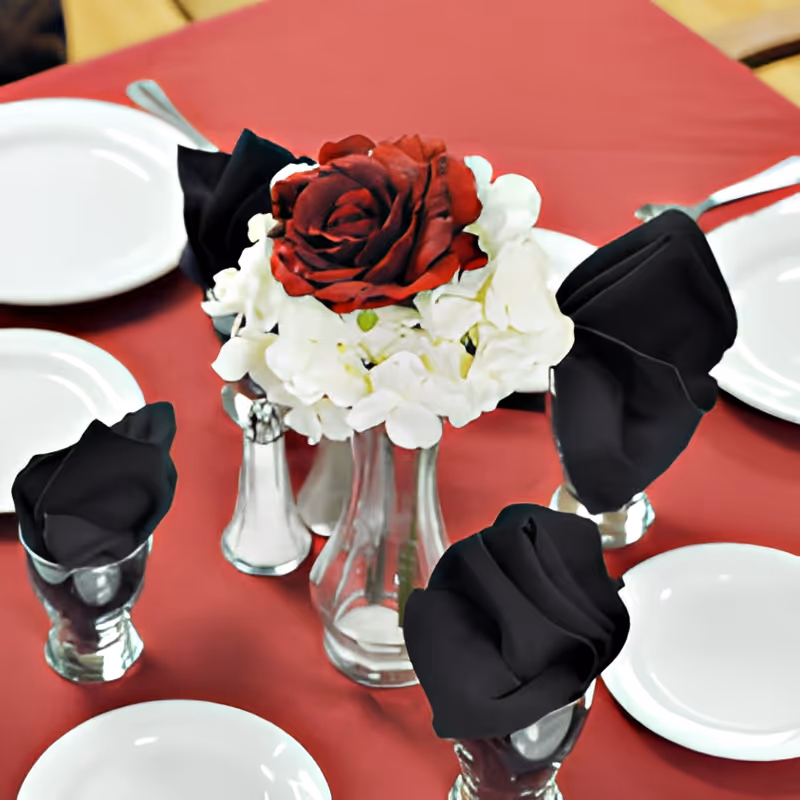 A dining table set with a red tablecloth, white plates, silver cutlery, and black folded napkins in silver napkin holders. In the center is a clear glass vase with a floral arrangement featuring a large red rose surrounded by white flowers.