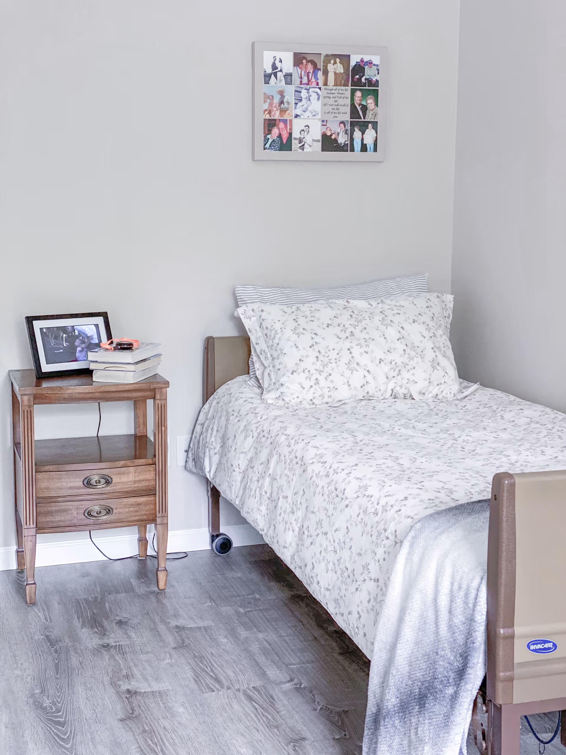A neatly made single bed with floral patterned bedding in a room with light gray walls and wood flooring. Next to the bed is a wooden nightstand with two drawers, holding a framed photo and a stack of books. Above the bed hangs a collage picture frame with multiple photos.