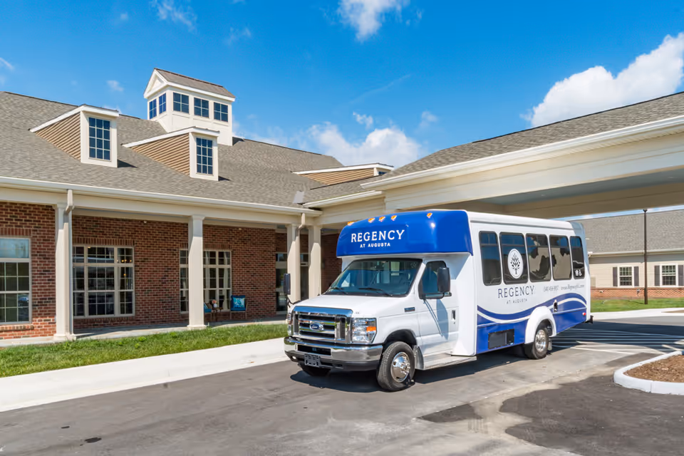 A white and blue shuttle bus with the text 'REGENCY AT AUGUSTA' parked in front of a brick building with large windows and a covered entrance under a blue sky with some clouds.