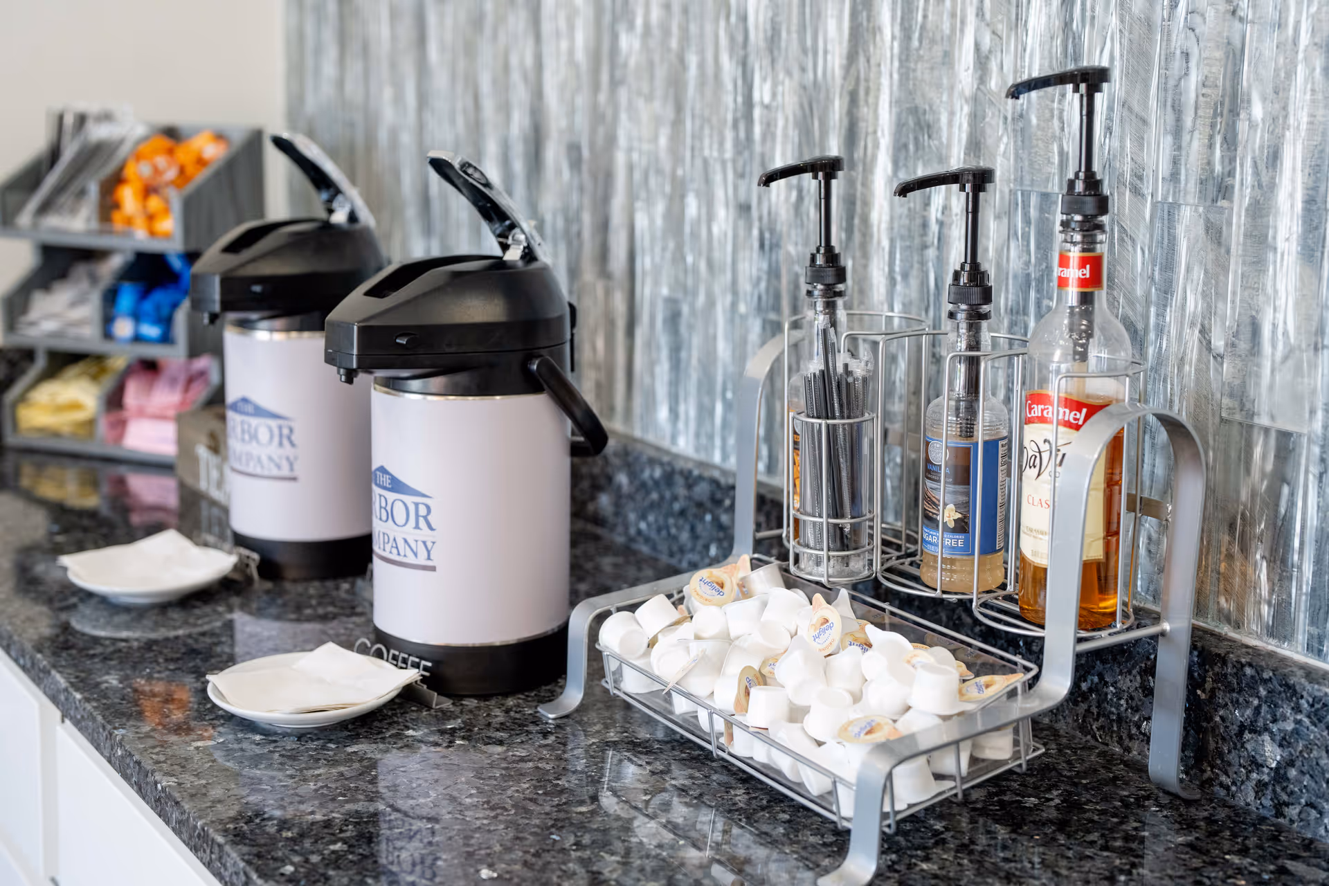 A coffee station with two airpot coffee dispensers labeled 'The Arbor Company', a tray filled with creamers, and three pump bottles of flavored syrups on a granite countertop with a textured silver backsplash.