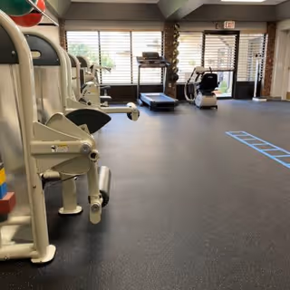 Interior view of a fitness room with exercise equipment including a treadmill, stationary bike, and weight machines. Large windows with blinds allow natural light to enter the room. The floor is covered with dark rubberized gym flooring and there is an exit door in the background.