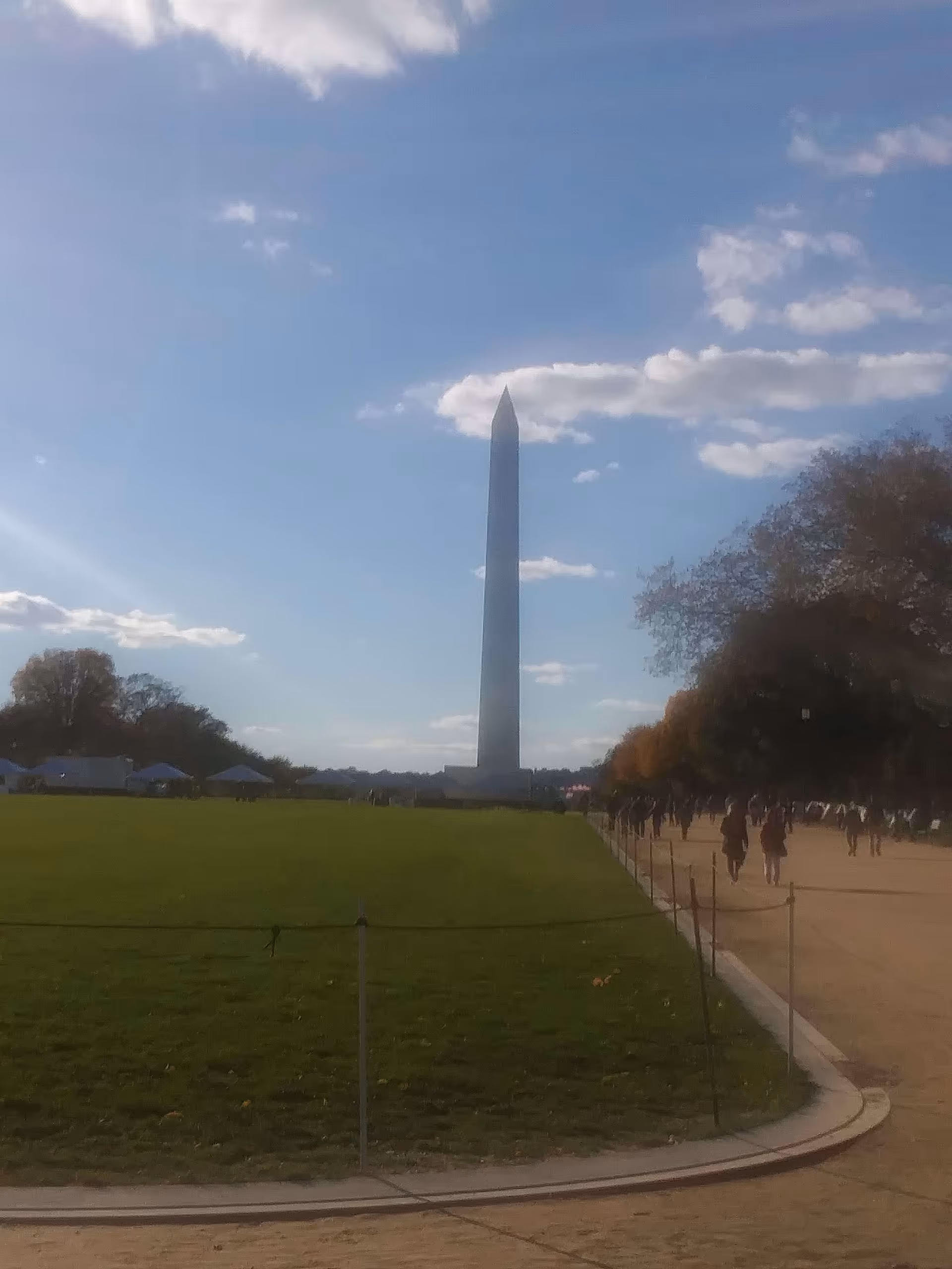 A tall, pointed obelisk monument standing in the center of a large grassy area with a clear blue sky and scattered clouds above. People are walking along a path to the right side of the monument, and trees line the path.