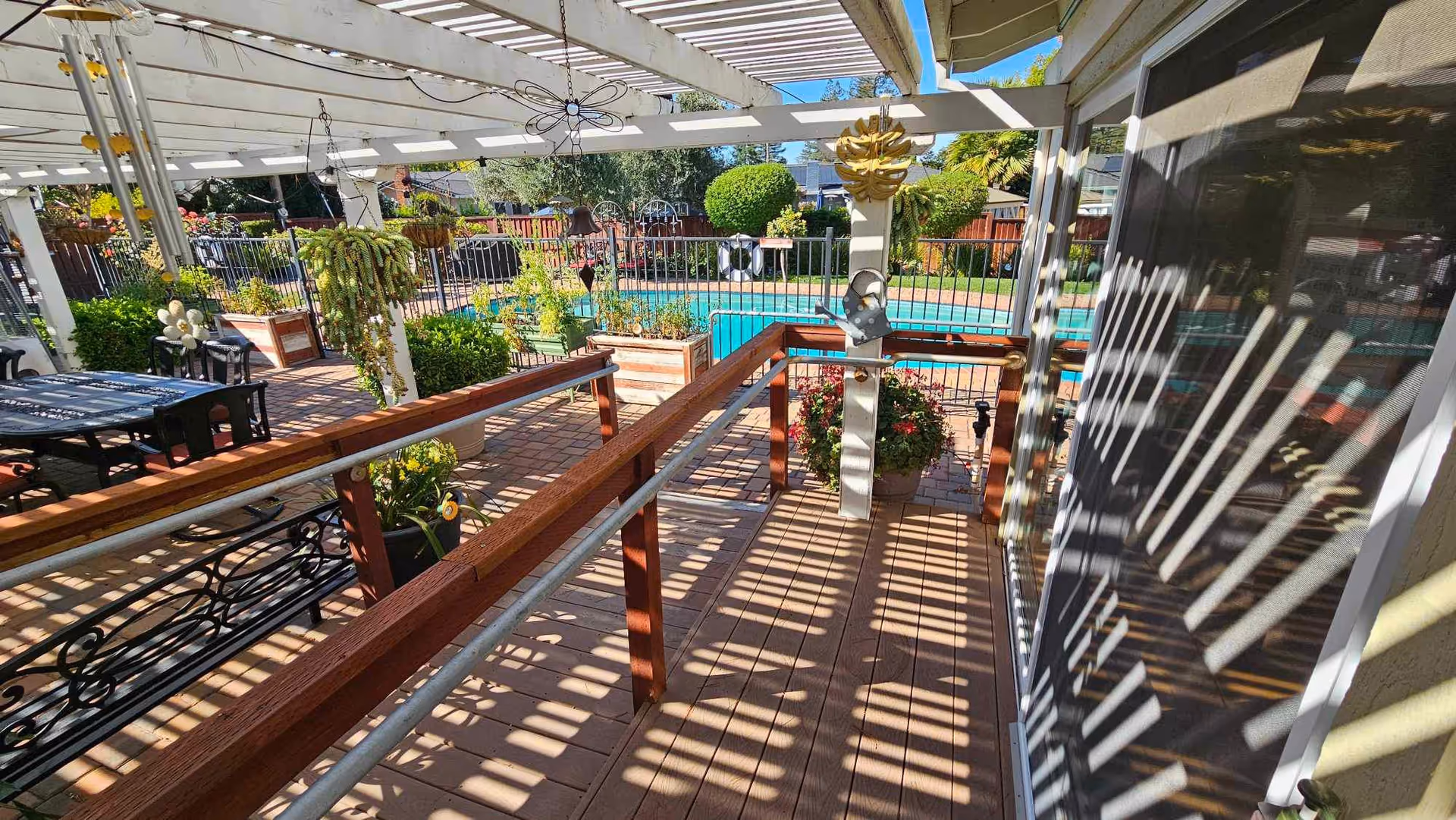 Outdoor patio area with wooden decking and a pergola casting striped shadows. There are hanging plants, potted flowers, and a wooden handrail along a ramp. In the background, a fenced swimming pool and well-maintained garden with bushes and trees are visible under a clear blue sky.