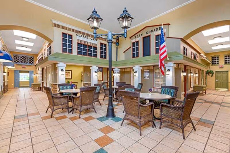A bright indoor commons area with round dining tables and wicker chairs arranged around a decorative streetlamp beneath mock storefront facades.
