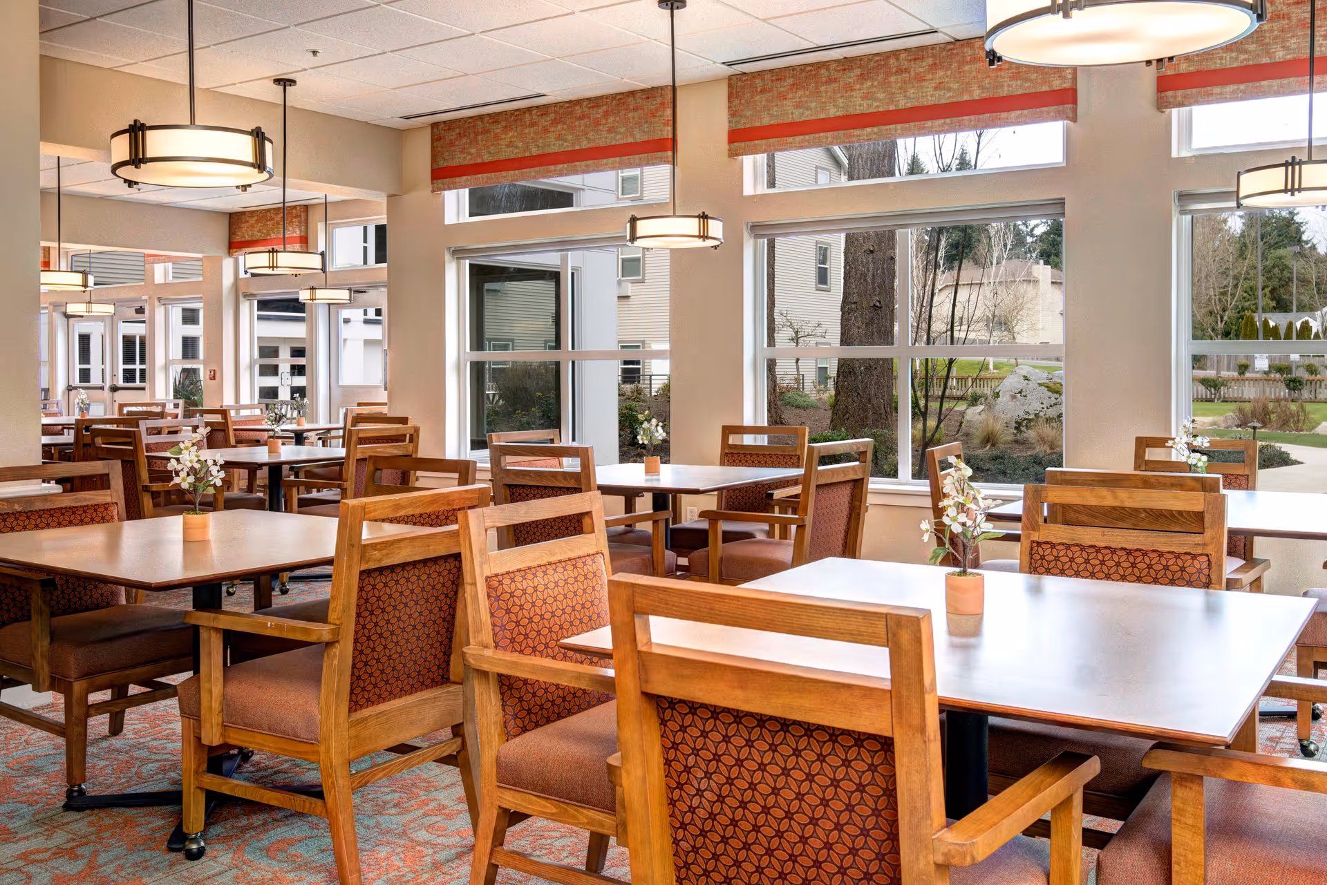 A bright dining room with wooden tables and chairs featuring patterned cushions. Small flower arrangements are placed on each table. Large windows allow natural light to fill the room and provide a view of the outdoor garden area. Modern pendant lights hang from the ceiling.