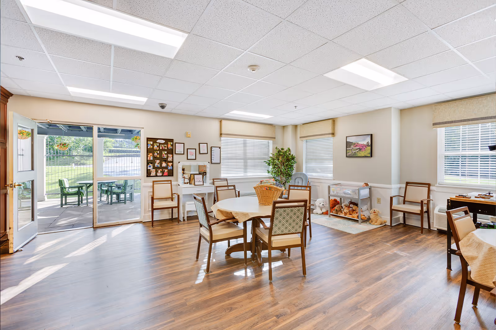 A bright and spacious common room with wooden flooring and several chairs around a round table covered with a beige tablecloth. The room has large windows with blinds, a small white vanity table with a mirror, and a basket on the table. There is a collection of stuffed animals in a clear storage bin against the wall, a potted plant, and framed pictures on the walls. A door and large windows open to an outdoor patio area with green chairs and a table.
