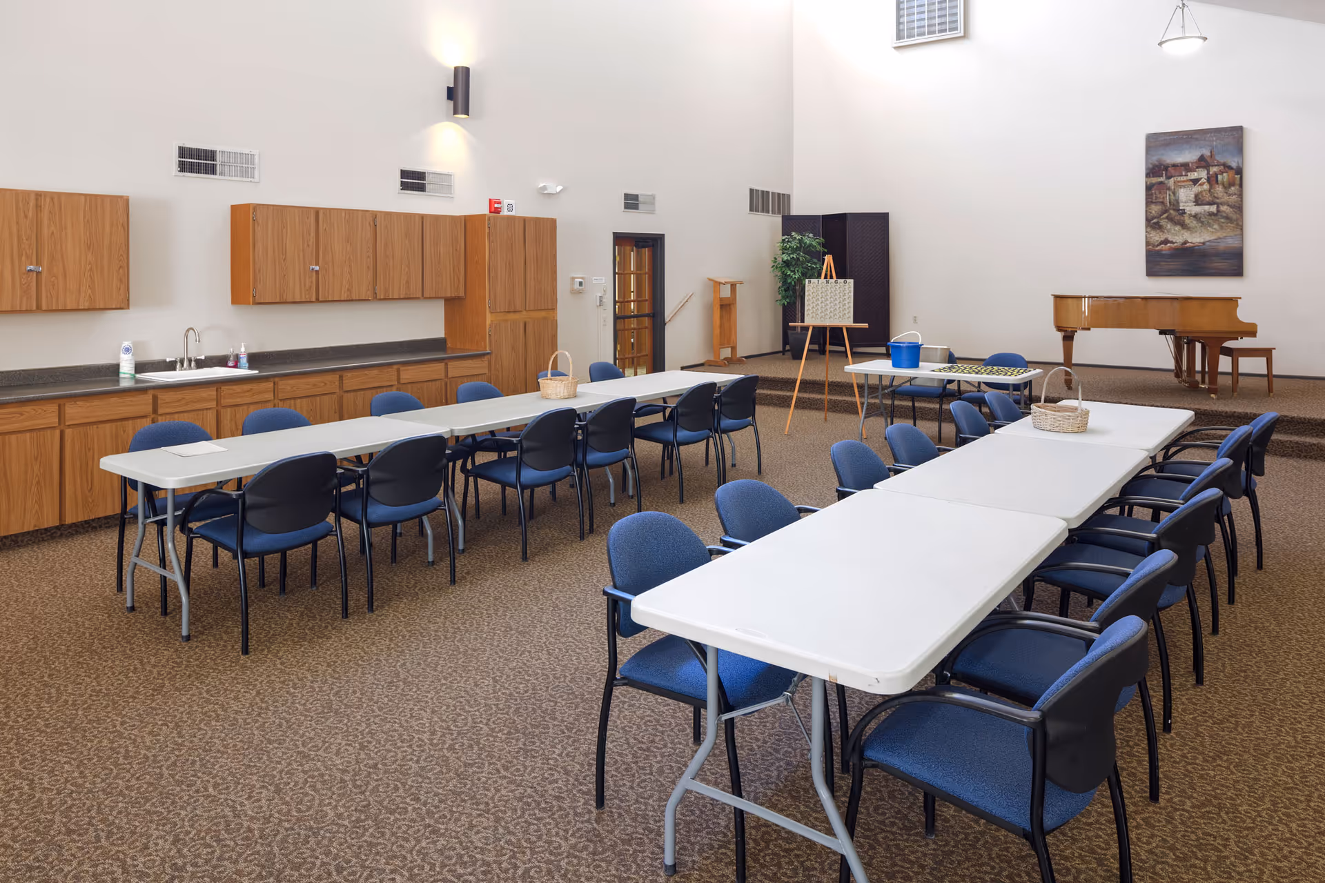 A spacious room with two long white tables arranged in a U-shape, surrounded by blue cushioned chairs. The room has wooden cabinets and a countertop with a sink along one wall. In the background, there is a small stage area with a grand piano, a painting on the wall, and an easel with a board. The carpet is patterned and the walls are light-colored with high ceilings.
