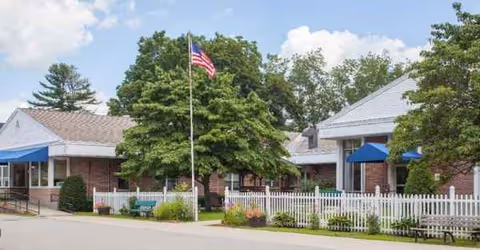 Exterior view of Alpine Healthcare Center with brick building, white fence, flag, and trees