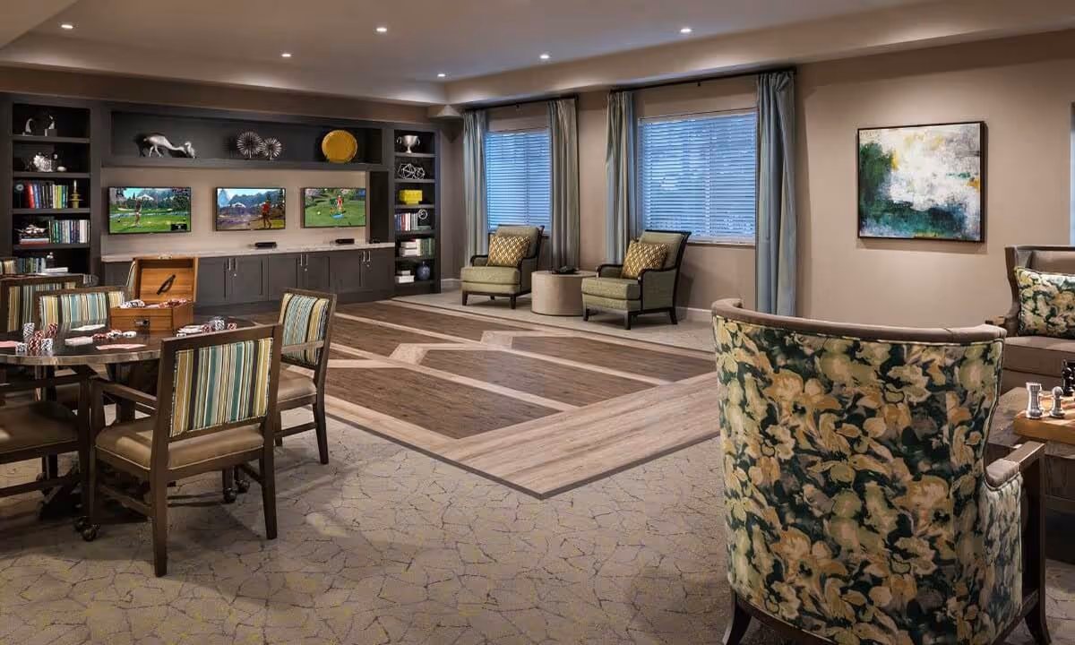 Spacious senior living community lounge with multiple seating areas, a game table, built-in shelving with three TVs, and large windows.