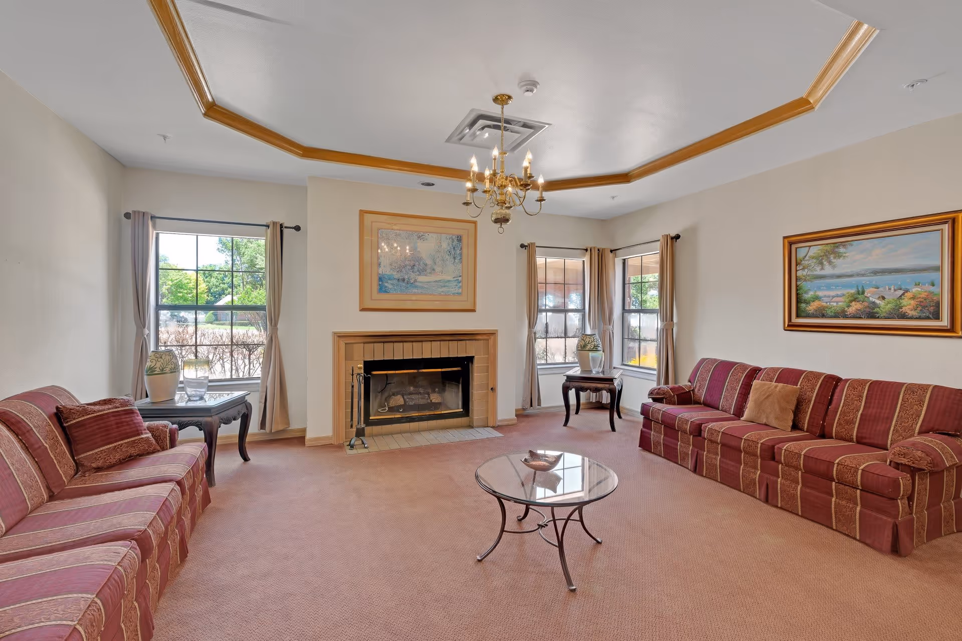 A cozy living room with two red patterned sofas facing each other, a glass coffee table in the center, and a fireplace with a framed painting above it. The room has beige walls, carpeted floor, two windows with beige curtains, and a chandelier hanging from the ceiling with wooden trim.