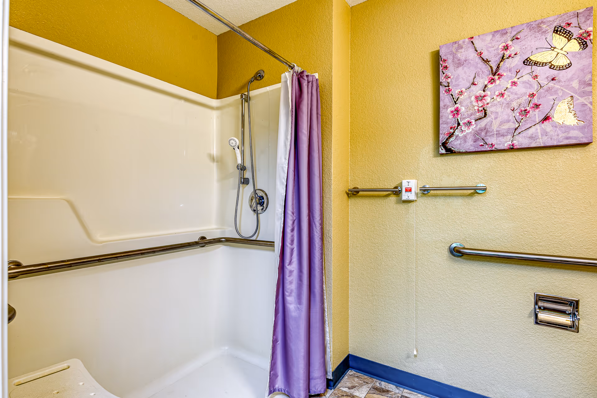 A bathroom shower area with yellow walls, a white shower stall equipped with a handheld showerhead, grab bars, and a purple shower curtain. There is a floral and butterfly artwork on the wall, along with a pull cord and a towel bar.