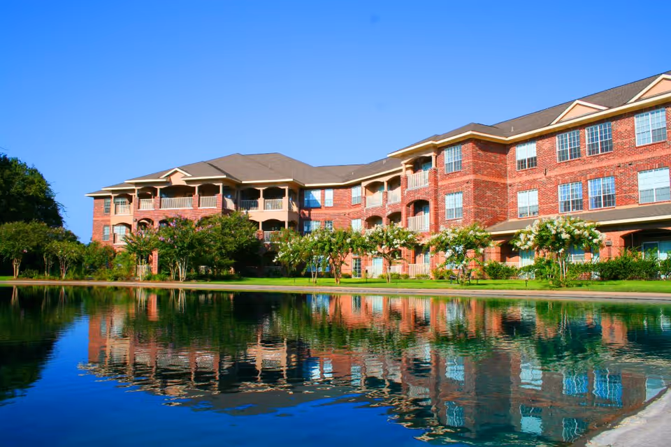 A large three-story red brick building with multiple windows and balconies, reflected in a calm pond in the foreground. The building is surrounded by green grass, trees, and shrubs under a clear blue sky.