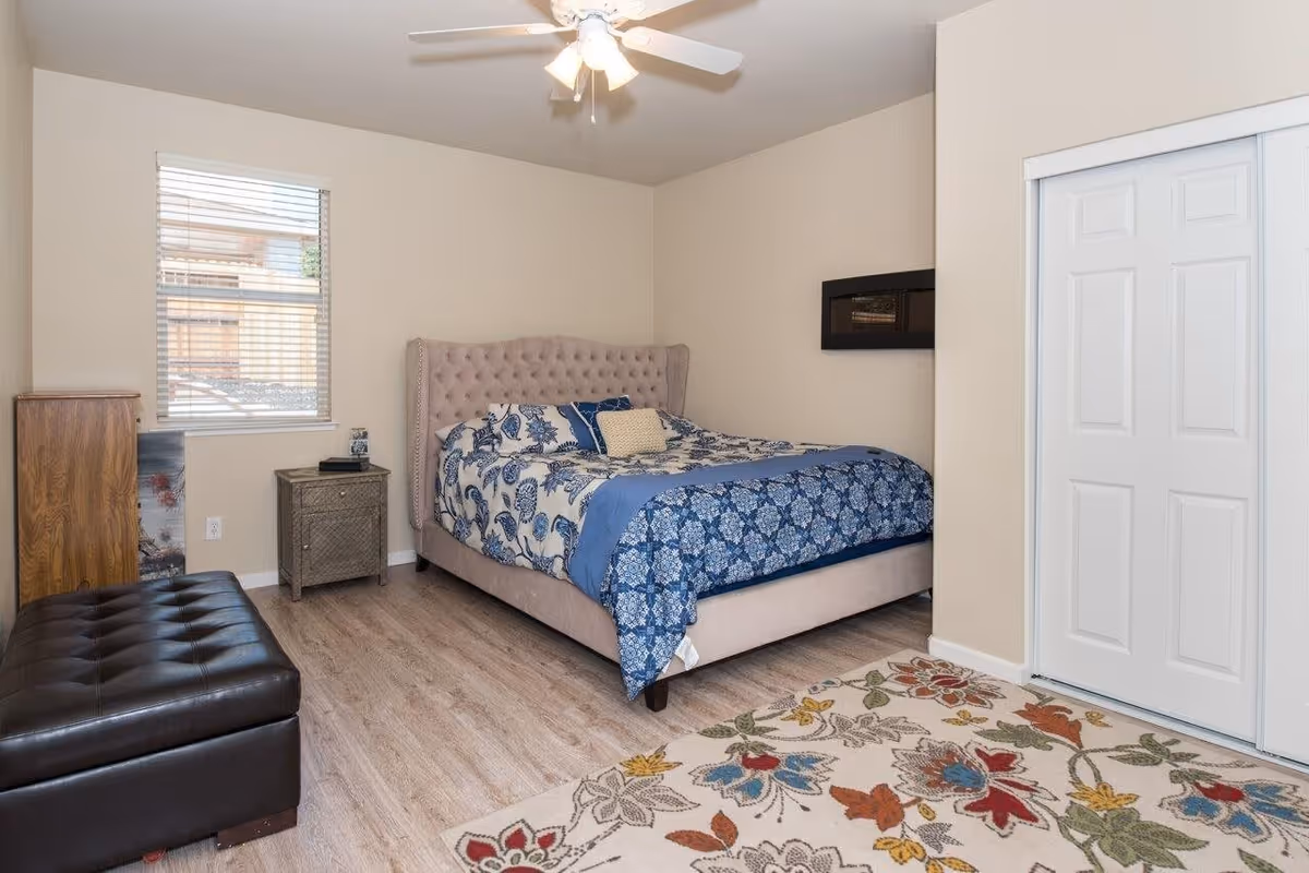 A bedroom with a beige tufted bed frame and blue floral bedding. The room has light wood flooring, a colorful floral area rug, a ceiling fan with lights, a window with blinds, a small nightstand with a lamp and clock, a wooden cabinet, and a dark brown leather bench at the foot of the bed. There is a white closet door on the right wall.