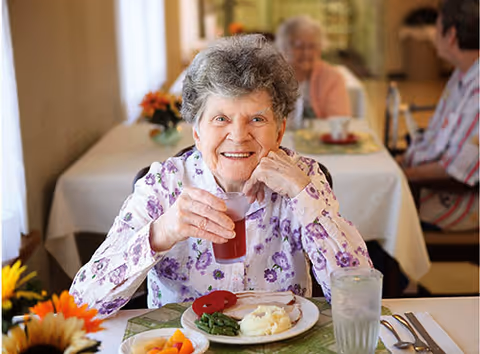 An elderly woman with gray curly hair wearing a floral blouse is sitting at a dining table holding a glass of red juice and smiling. On the table in front of her is a plate with mashed potatoes, green beans, and a slice of meat, along with a glass of water. In the background, two other elderly individuals are seated at another table in a dining room setting.