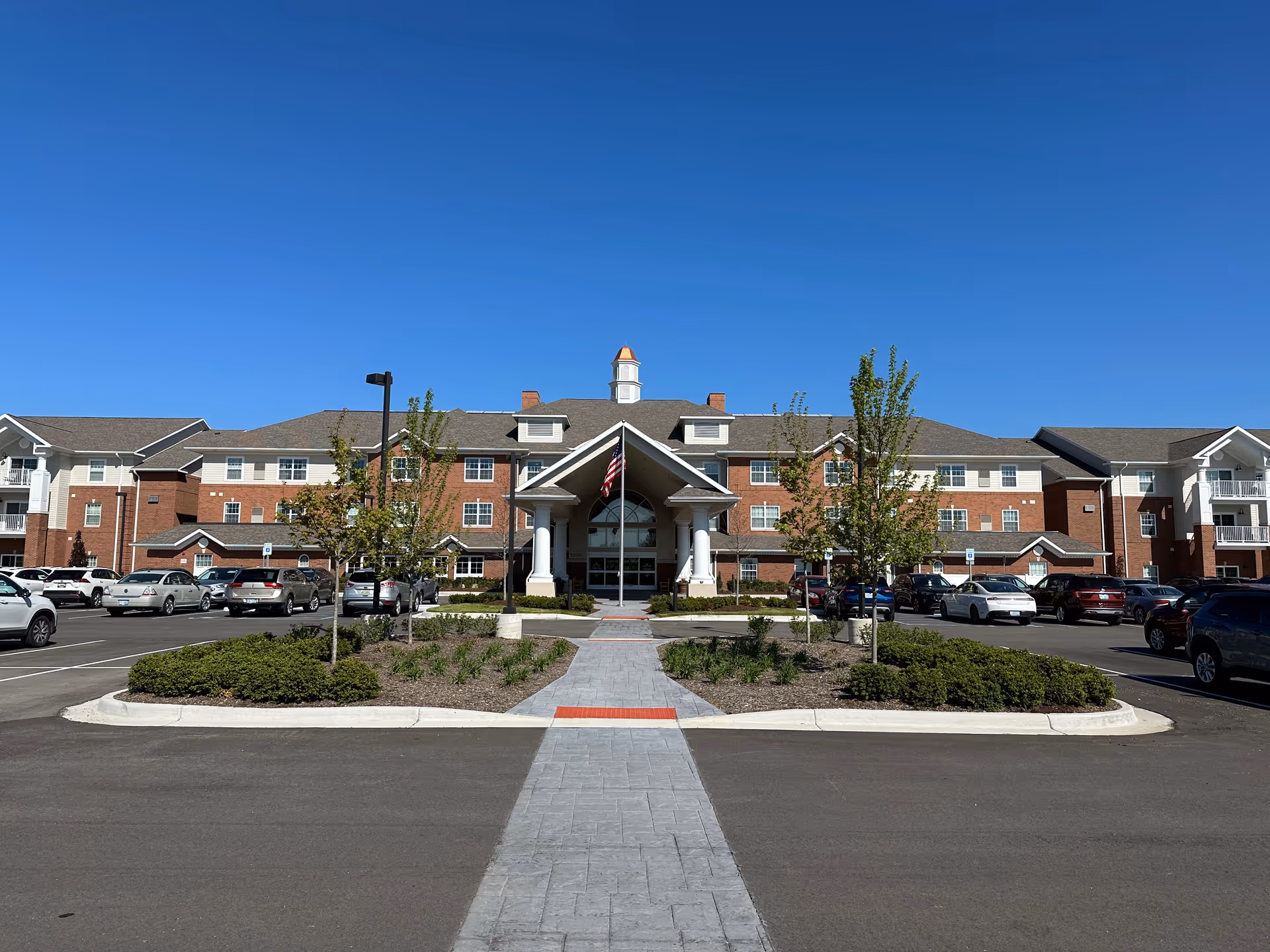 Front exterior view of Willow Pines Retirement Community building with a clear blue sky, a paved walkway leading to the entrance, landscaped greenery, and parked cars on both sides.