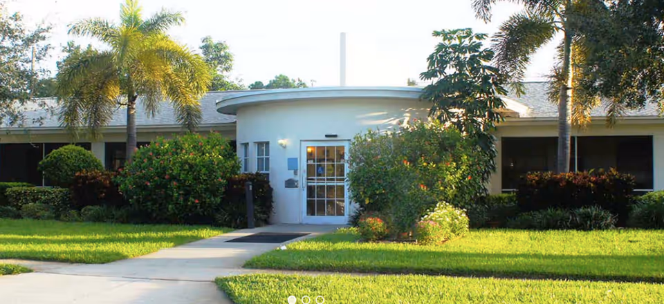 Front exterior view of Abbey Rehabilitation and Nursing Center showing a single-story building with a rounded entrance, glass door, surrounded by green bushes, palm trees, and a well-maintained lawn with a concrete walkway leading to the entrance.