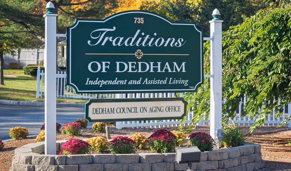 Outdoor sign for Traditions of Dedham, an independent and assisted living facility, located at 735. Below it is a smaller sign for the Dedham Council on Aging Office. The signs are mounted on white posts surrounded by a flower bed with colorful flowers and greenery.