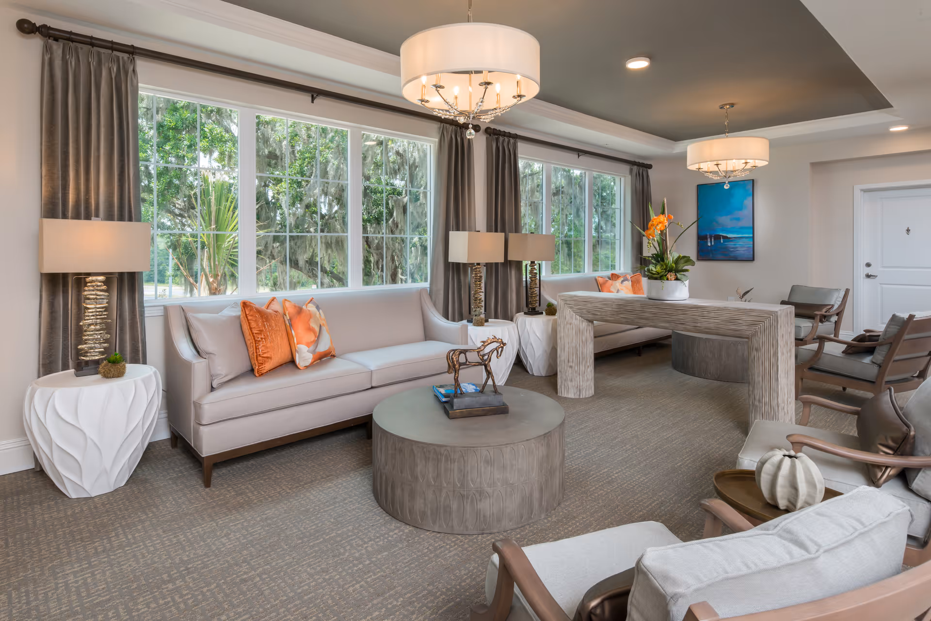 A bright and spacious living room with large windows showing green trees outside. The room features two light gray sofas with orange and patterned throw pillows, two modern table lamps on white side tables, a round gray coffee table with a decorative horse sculpture, and a rectangular wooden console table with a flower arrangement. There are also several cushioned chairs, a blue painting on the wall, and two elegant ceiling light fixtures.