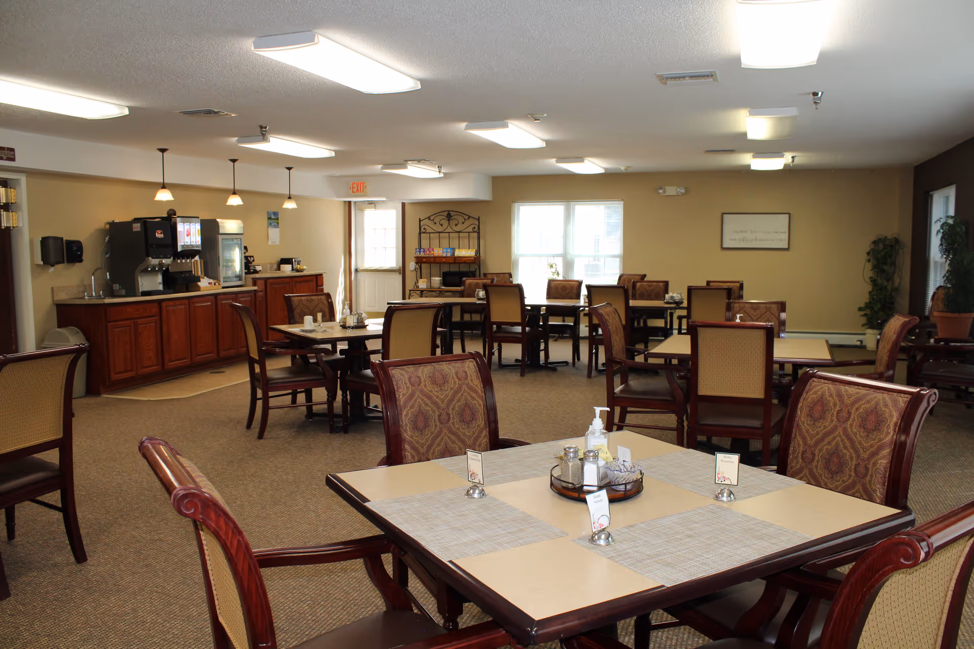 Bright communal dining room with multiple tables and chairs and a beverage/serving station along the back wall.