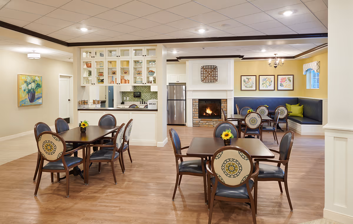 A cozy dining area in a senior living facility featuring several wooden tables with decorative chairs. The room has a warm ambiance with a fireplace, a kitchen area with a refrigerator and cabinets, and a built-in bench seating with cushions along one wall. The walls are painted yellow and beige, and there are framed floral artworks and a painting of flowers on the walls. The floor is wooden, and the ceiling has recessed lighting and a chandelier.