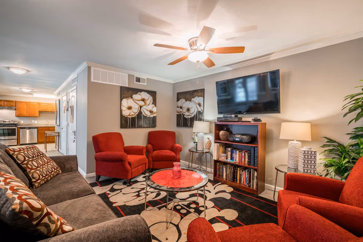 A cozy living room with a gray sofa adorned with patterned pillows, three red armchairs, a round glass coffee table with a red centerpiece, and a patterned rug. The room features two floral paintings on the wall, a ceiling fan with a light, a wooden bookshelf filled with books and a stereo system, a flat-screen TV mounted above the bookshelf, two side tables with lamps, and a large green plant in the corner. The kitchen area with wooden cabinets and stainless steel appliances is visible in the background.