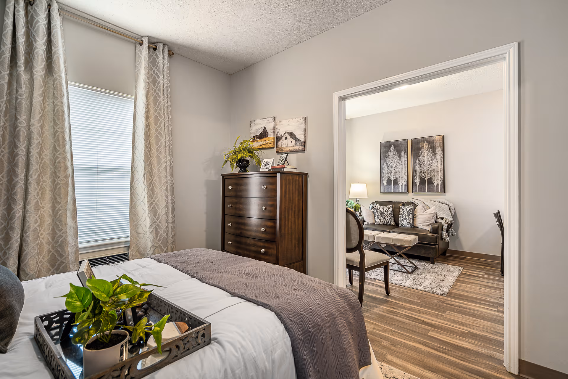 A cozy bedroom with a bed covered in white and gray bedding, a decorative tray with a small potted plant on the bed, a wooden dresser with framed pictures and a plant on top, and a window with patterned curtains. Through an open doorway, a living room is visible with a dark sofa, patterned cushions, a lamp, a chair, and wall art featuring tree designs.