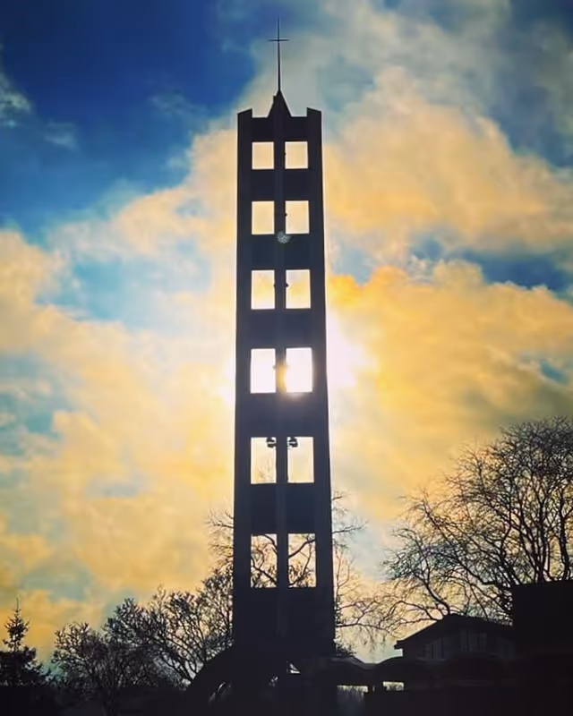 Tall bell tower with a cross on top silhouetted against a bright sky with clouds and the sun shining through the tower's openings. Trees and part of a building are visible at the base of the tower.