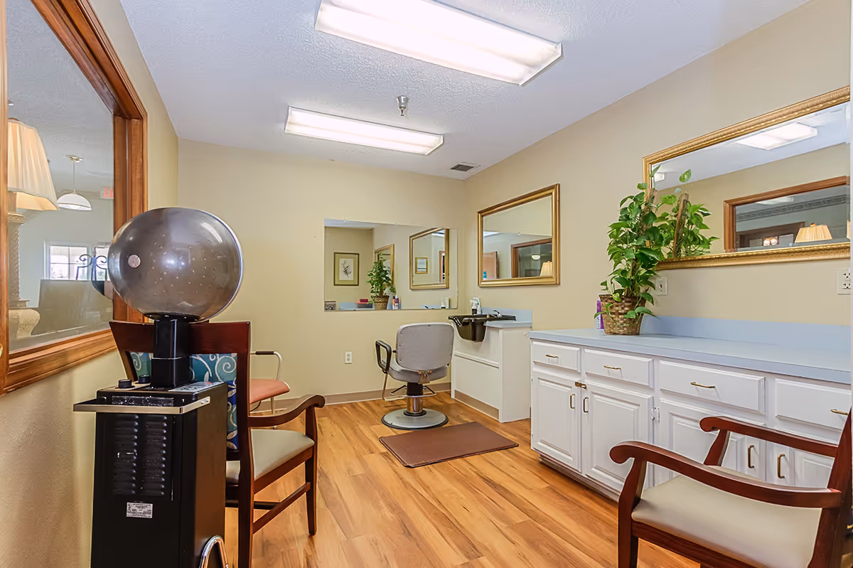 Interior view of a senior living facility's hair salon area with a hair dryer chair, a salon chair in front of a mirror, wooden chairs, white cabinets with a countertop, a potted plant, and bright overhead lighting.