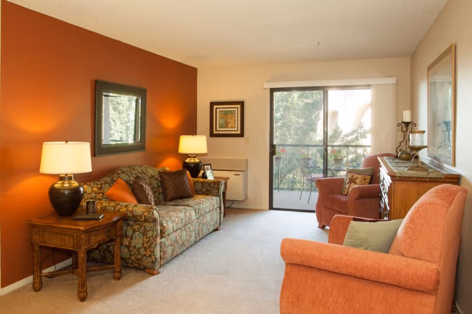 Living room with a patterned sofa, two upholstered armchairs, side tables with lamps, and a sliding glass door to a balcony.