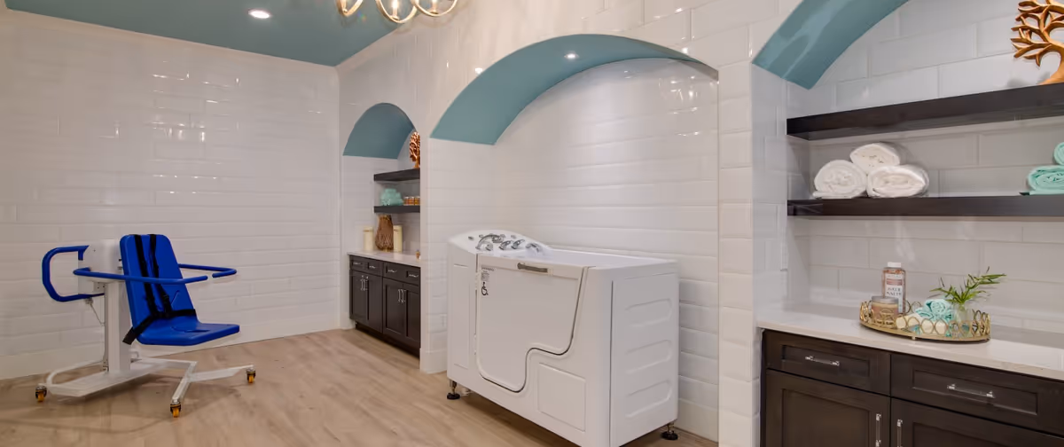 A clean and modern bathing room featuring a white accessible walk-in bathtub with control knobs, a blue assisted bathing chair with safety straps, white tiled walls with arched alcoves painted light blue, dark wood cabinets with rolled towels and bath supplies on open shelves, and light wood flooring.