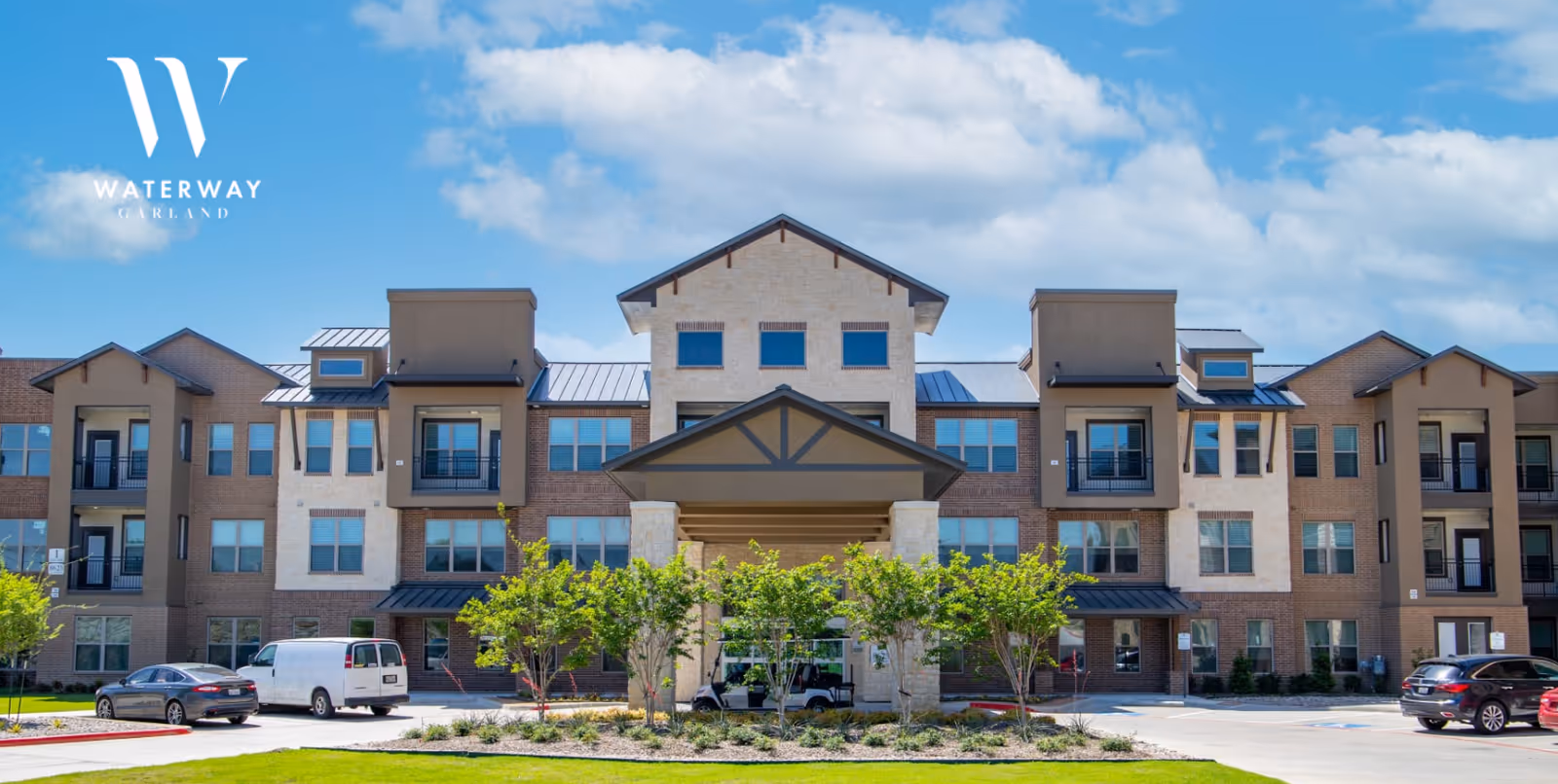 Front entrance of a multi-story senior living building with a covered porte-cochere, landscaping, and parked vehicles under a blue sky.