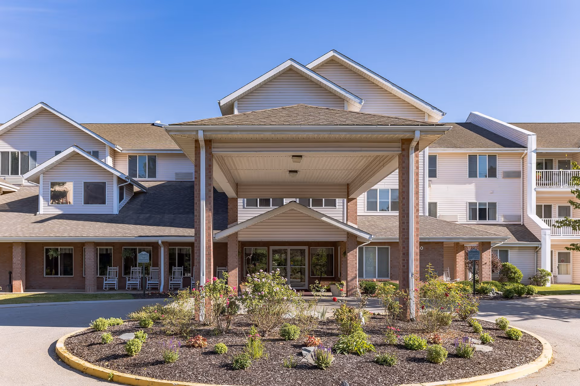 Front exterior view of Provincial Bethel Park facility with a covered entrance, multiple windows, and a landscaped circular flower bed in front. The building has a combination of brick and siding with a clear blue sky above.