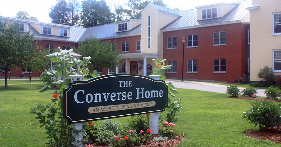 Exterior view of The Converse Home, an assisted living community, showing a large brick and yellow building with multiple windows and a metal roof. In the foreground, there is a green sign with white and gold lettering surrounded by plants and flowers on a well-maintained lawn.