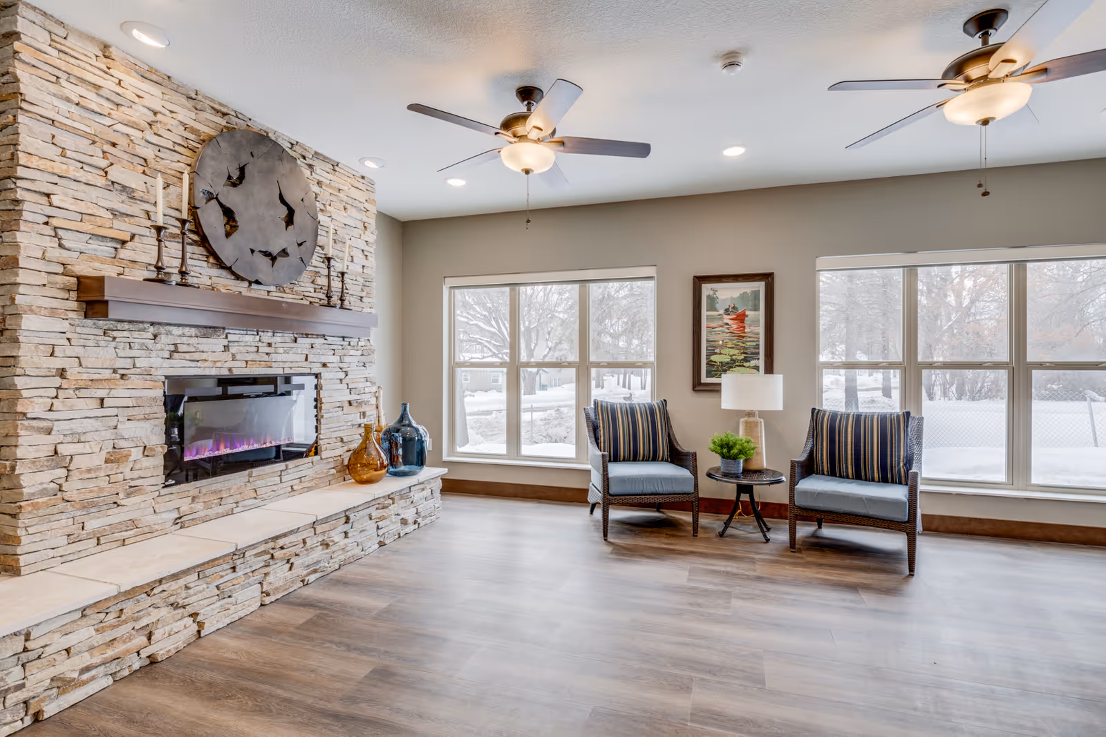 A cozy living room with a stone fireplace featuring a modern electric insert and a large decorative clock above it. Two armchairs with striped cushions are positioned near a small round table with a lamp and a small plant. Large windows let in natural light and show a snowy outdoor scene. The room has wood flooring and ceiling fans with lights.