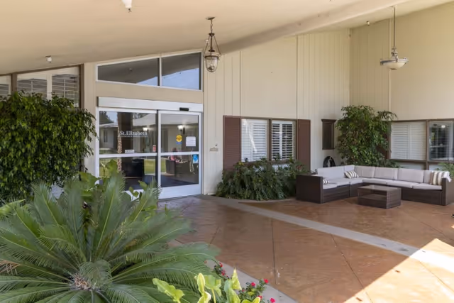 Covered entrance to St. Elizabeth Healthcare & Rehab showing glass doors, potted plants and a wicker seating area under a canopy.