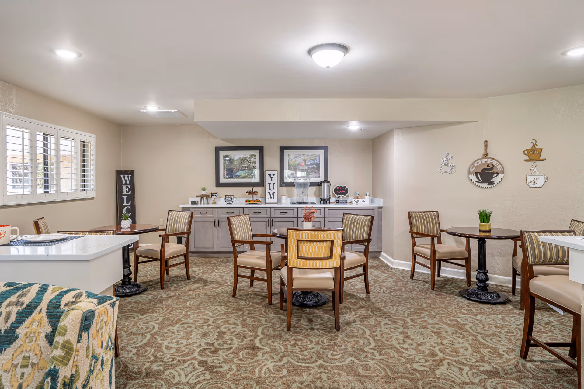 A cozy dining area with several round tables and wooden chairs with cushioned seats. The room has beige walls and patterned carpet flooring. On the back wall, there is a counter with cabinets underneath, decorated with framed pictures, a coffee station, and small decorative items. Wall decorations include coffee-themed signs. A window with white shutters is on the left side, and the room is well-lit with ceiling lights.