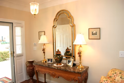 Interior view of a well-lit entryway featuring a decorative wooden console table with a marble top. On the table are two ornate lamps with white shades, a large decorative vase, and three framed photographs. Above the table hangs a large, ornate gold-framed mirror reflecting a white door. To the left is a glass door with white blinds, and to the right is a floral-patterned armchair partially visible.