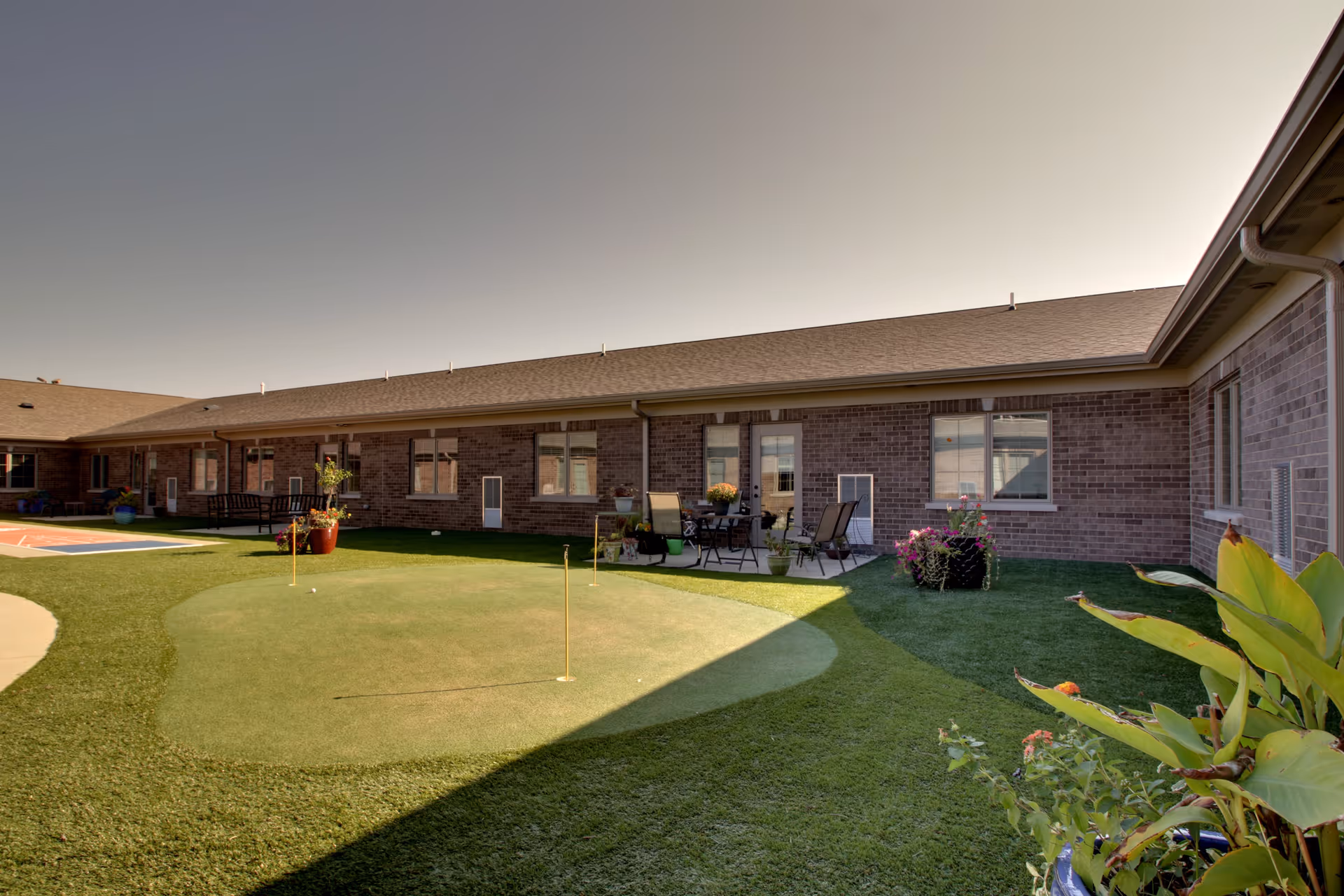 Outdoor courtyard area of a senior living facility with a small putting green, patio furniture, potted plants, and a brick building surrounding the space under a clear sky.