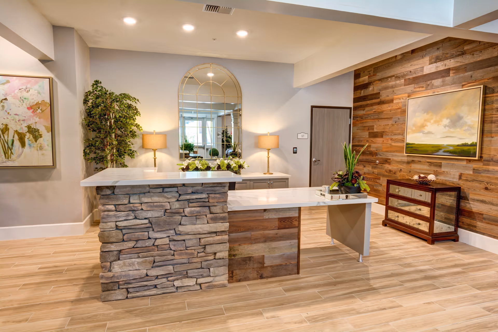 A reception area with a stone and wood front desk, two table lamps, a large decorative mirror, a potted plant, and framed artwork on the walls. The floor is wood-patterned tile and there is a wooden accent wall with a landscape painting and a small mirrored cabinet.