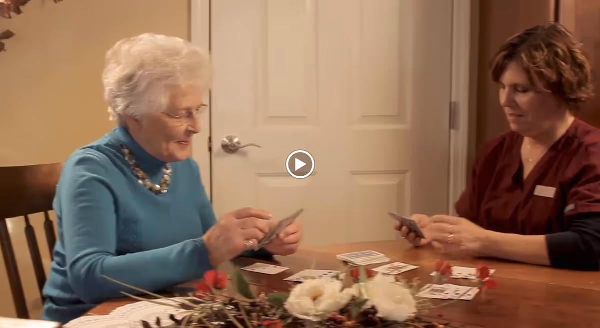An elderly woman and a caregiver sitting at a wooden table playing cards together in a cozy indoor setting. The table has a floral centerpiece and a lace doily. Both individuals appear engaged and content.